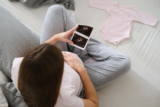 A young pregnant woman gently holds an ultrasound picture of her baby in her hands while sitting on the bed in the bedroom. Maternity concept.