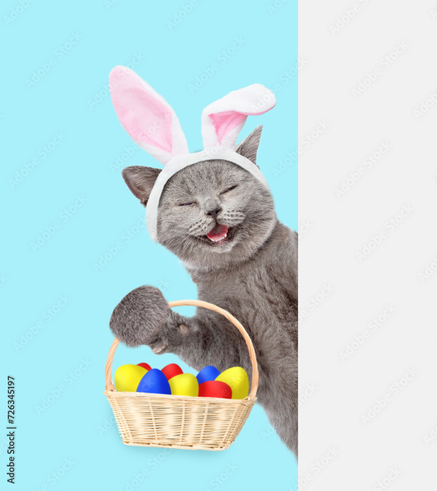 Aufkleber happy kitten wearing easter rabbits ears holds basket with colorful easter eggs and looks from behin