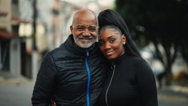 Middle-aged father and teen daughter posing together for camera standing outside in urban environment during drizzle rain. South American hispanic people of African descent