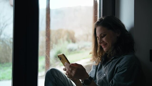 A young cheerful Caucasian woman uses her phone while sitting by the window