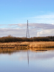 Electricity pylon reflected in a wetland pond with a blue sky background