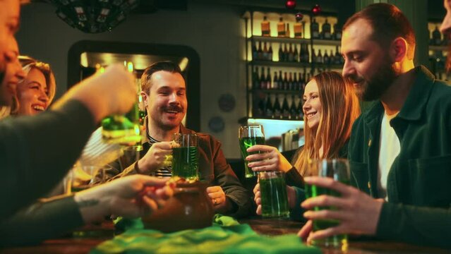 Cheerful young people, friends meeting at pub, clinking green beer glasses, talking, laughing, saying toasts. Celebration of St Patrick's Day. Concept of leisure time, holiday, fun and joy, traditions