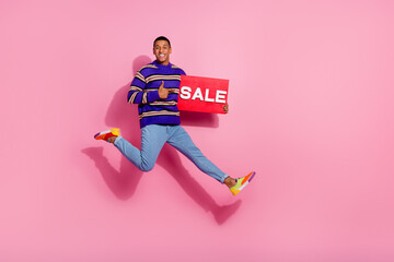 Full length photo of good mood confident man wear sweater jumping thumb up holding sale board empty space isolated pink color background
