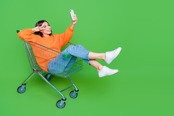 Full length photo of funky person inside pushcart hold smart phone make selfie show v-sign empty space isolated on green color background