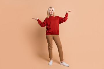 Full length photo of cheerful excited woman wear red sweater comparing arms empty space isolated beige color background