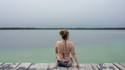 woman in bikini looking at green lagoon sitting on wooden pier