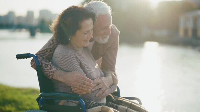Husband walking with wife in wheelchair. Senior family couple in city park. Old man hugging woman showing love care, they talking smiling. Living with physical disability, long love concept.
