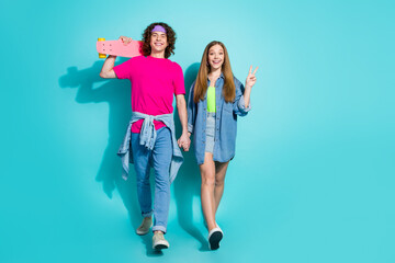 Fototapeta premium Full length photo of hipster guy with headband stroll with girlfriend teenager show v sign holding skate isolated on cyan color background