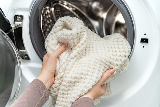 Woman putting white wool sweater into the drum of a washing machine in laundry room. 