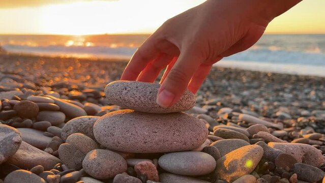 4K horizontal video. Woman builds pyramid of light stones on pebble beach. Setting sun, sea waves, coast, sunset.  Ocean shore zen. Concept of calm, yoga, travel, balance, meditation