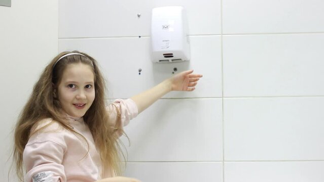 A little teenage girl dries her hands with air using an electric dryer in the bathroom or toilet. Hygiene concept
