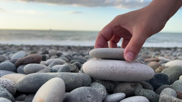 Female hand builds pyramid of gray stones on pebble beach with sea waves. Concept of spa, calmness, yoga, self-improvement, construction, games, travel. White woman experiences zen on ocean shore