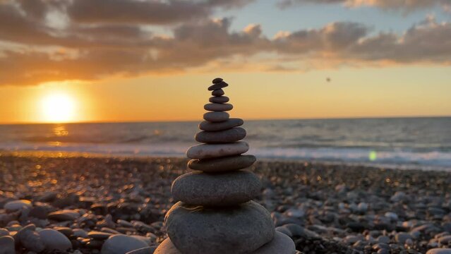 Camera rotates around pyramid of small stones on pebble beach against background of setting sun, waves and coast on sunset. Ocean view. Zen of seashore. Concept of calm, yoga, travel, tourism, life