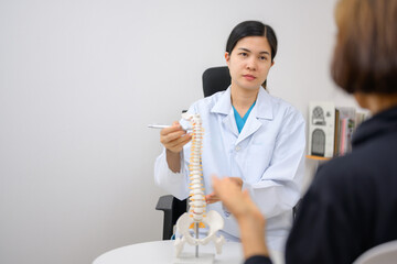 An orthopedic surgeon or therapist is showing a spinal model and explaining to a female patient her...