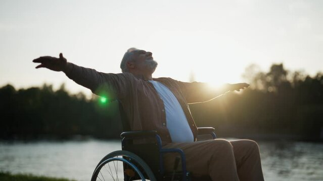 Senior man in wheelchair enjoys freedom. Paralysis old male with special need spreading arms on sides smiling breathing fresh air walking in city park. Living with physical disability concept.