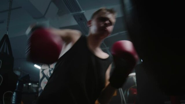 African American man in boxing gloves exercises with trainer in dark gym before fighting competition. Boxer hits punching bag while practicing in boxing gym. Physical activity and intensive workout.