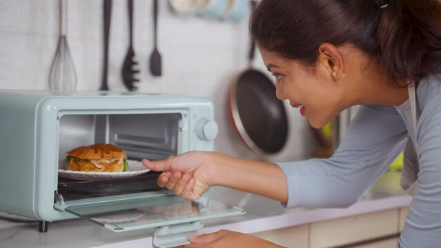 Indian woman cooking burger by placing it on microwave oven at kitchen - concept of healthy eating, domestic lifestyle and technology