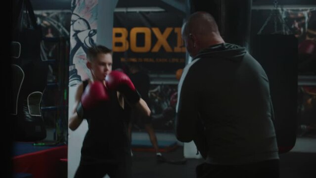 Focused young fighter hits punching bag while training in dark boxing gym. Athletic teenager in boxing gloves exercises with coach before fighting tournament. Physical activity and intensive workout.