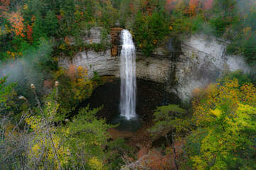 Fototapeta na wymiar Falls Creek Falls is surrounded by vibrant fall colors and a misty morning haze. The waterfall cascades from a rocky cliff into a dark pool below. Photographed at Falls Creek Falls State Park in TN