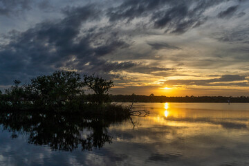 The setting sun is reflected in the Tomoka River, silhouetting a mangrove island. The trees and clouds are reflected in the water in this dramatic landscape. Photographed in Tomoka State Park, FL.