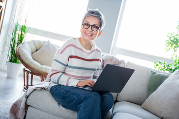 Photo of positive grandmother sitting comfy couch use wireless laptop modern spacious apartment...