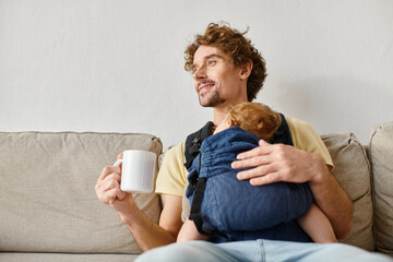 cheerful father with infant son in baby carrier holding cup of tea in living room, fatherhood