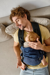 top view of pleased father sitting on couch with infant son in baby carrier, fatherhood and love