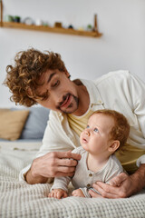 happy man with curly hair embracing his baby boy while lying together on bed, nurturing and love