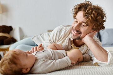 joyful father with curly hair and beard looking at his baby son while lying together on bed, love
