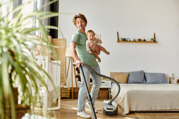 man multitasking housework and childcare, curly father vacuuming apartment with infant boy in arms - obrazy, fototapety, plakaty