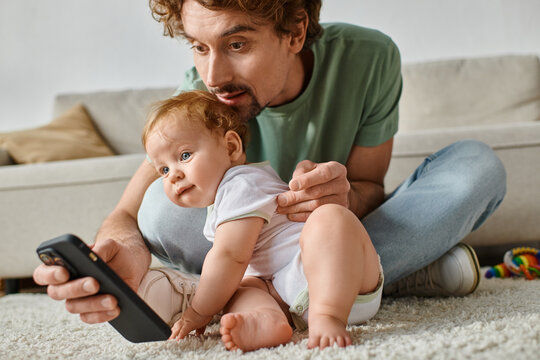 cheerful single father using smartphone while sitting with infant baby boy on carpet at home
