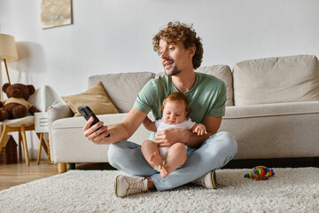 happy single father using smartphone while sitting with infant baby boy on carpet near rattle