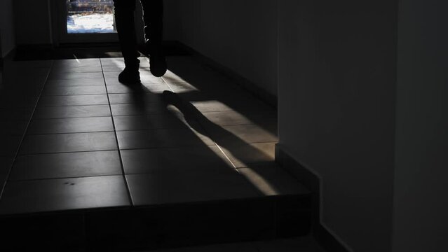Сlose-up of man's legs walking towards the exit of the room along narrow corridor in backlight. Backlit man walks across the room and opens the door and goes outside. Silhouette of a man.