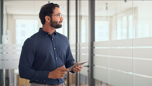 Smiling professional Indian businessman using tab computer working standing in office. Busy professional business man entrepreneur holding digital tablet tech device working, looking at pad.
