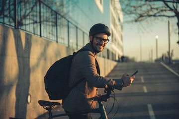 A portrait of a happy businessman with a backpack using a smartphone and leaning on handlebars of a...