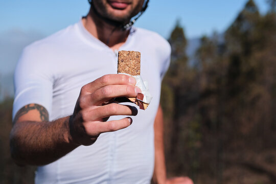 Male cyclist wearing cycling kit and helmet is holding energy snack for endurance during bicycle training. Sport nutrition. Sport snack for cyclist. Granola energy bar.