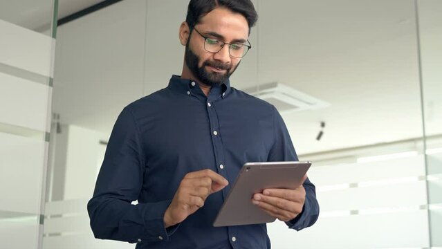 Busy Indian business man project manager looking at digital tablet walking in office. Professional businessman entrepreneur going in hallway holding tab working using tab computer.
