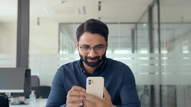 Busy Indian business man worker using cell phone at work in office. Professional businessman entrepreneur working on cellphone, smiling male employee executive holding smartphone checking mobile apps.