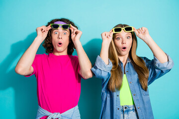 Portrait of impressed reaction shocked couple teenagers in bright clothing take off their sunglasses isolated on blue color background