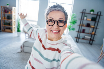 Photo of friendly positive grandmother toothy smile take selfie arm demonstrate modern house indoors