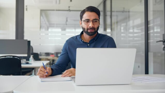 Busy Indian business man manager wearing earbuds having hybrid virtual conference meeting, watching online webinar, writing notes, communicating online working on laptop computer in office.