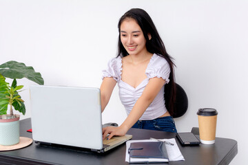 A young, cheerful female college student focusing on her laptop while studying at a neatly organized desk. At a white background.