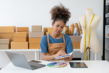 Young African woman running an online store, startup, small business, SME, using a smartphone or tablet, works with receipt boxes and checks online orders to prepare to pack boxes to sell to customer