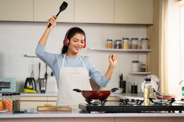 Joyful Dancing Indian woman cooking by listening songs on wireless headphones at kitchen - concept of weekend fun, food preparation and entertainment.