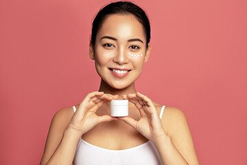 Cheerful woman presenting a cream jar with smears of product on her face against a pink background.