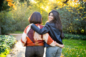 Happy mother and daughter walking hugging in a park and the daughter looks back smiling at the...
