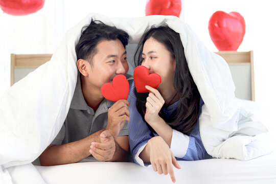 Happy cute Asian couple holding red heart during lying down on bed together, covering with white blanket. Romantic lover celebrating anniversary on Valentine Day in beautifully decorative home bedroom