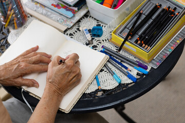 elderly wrinkled hands holding a pencil and writing in a notebook with drawing supplies on table