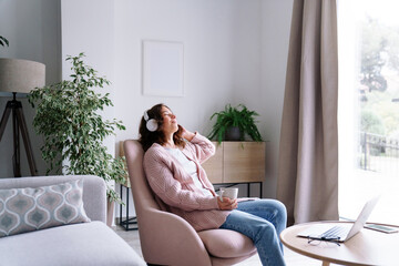 Young woman relaxing at home. Girl in headphones listen to music in cozy living room, using audio healing apps for mental health. Female wellness rituals and self-care lifestyle, calming sound therapy