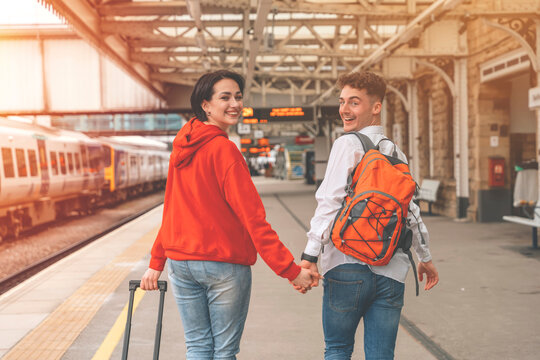 Happy loving couple with luggage and backpack walking along railway platform as they are ready to travel on holiday. Man and woman holding  hands,  looking back and smiling. Travel by train concept
